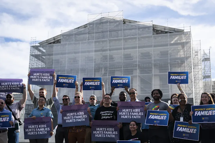 People hold signs outside a tall building under construction protesting conversion therapy.