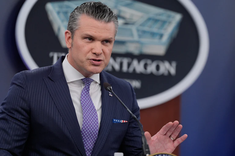A man in a navy suit and purple tie gestures as he speaks at a lectern.