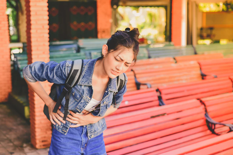A young woman wearing a denim jacket and a backpack holds onto her stomach in pain.
