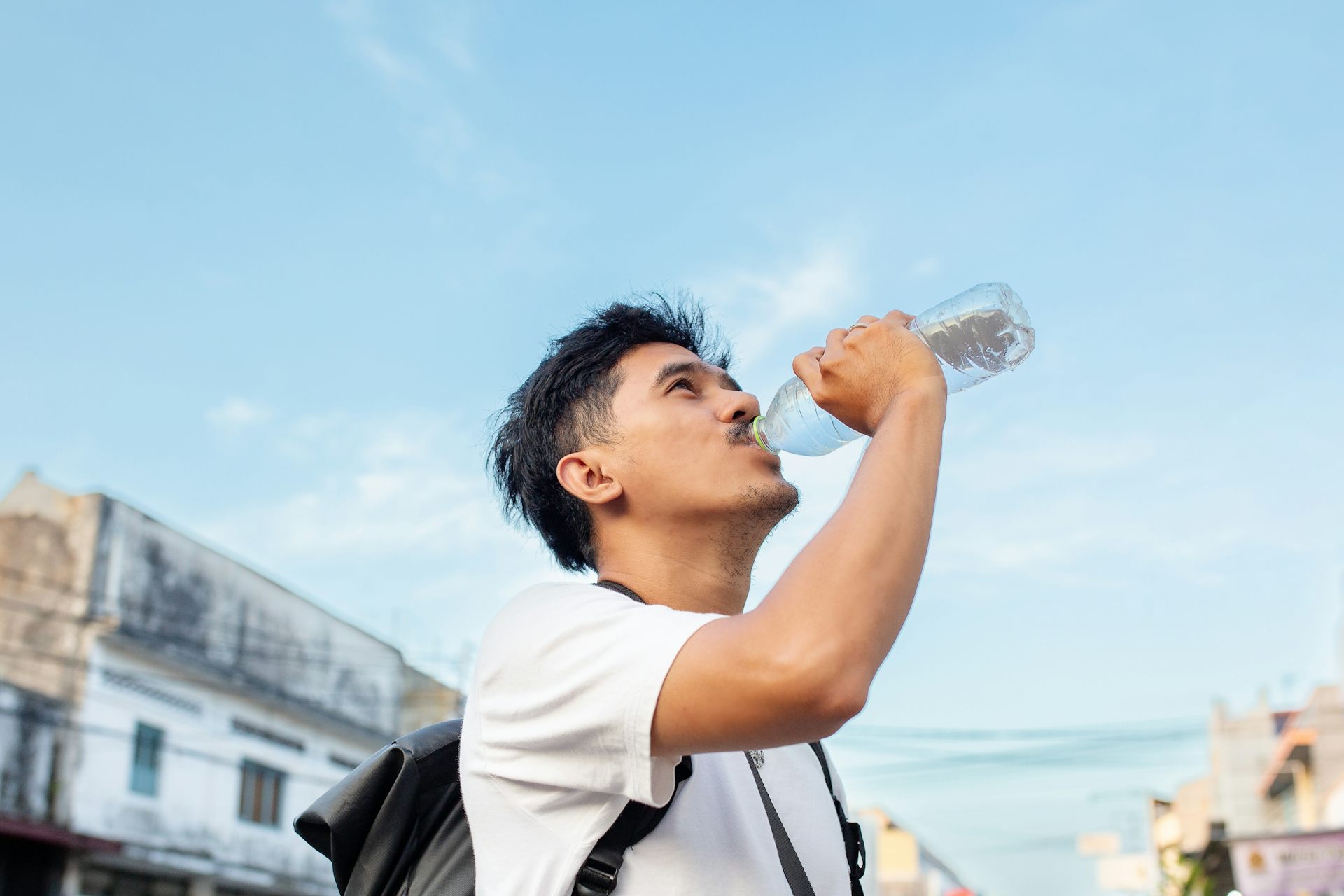 A young man wearing a backpack drinks water from a plastic bottle.