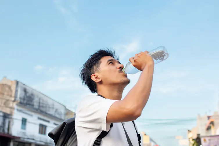A young man wearing a backpack drinks water from a plastic bottle.