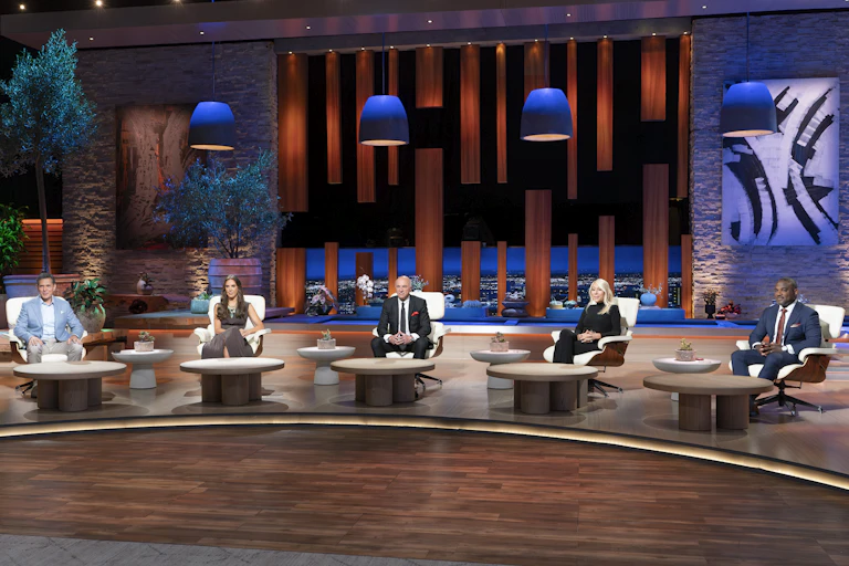 3 men and 2 women in formal clothes sit in chairs in front of tables on a big stage