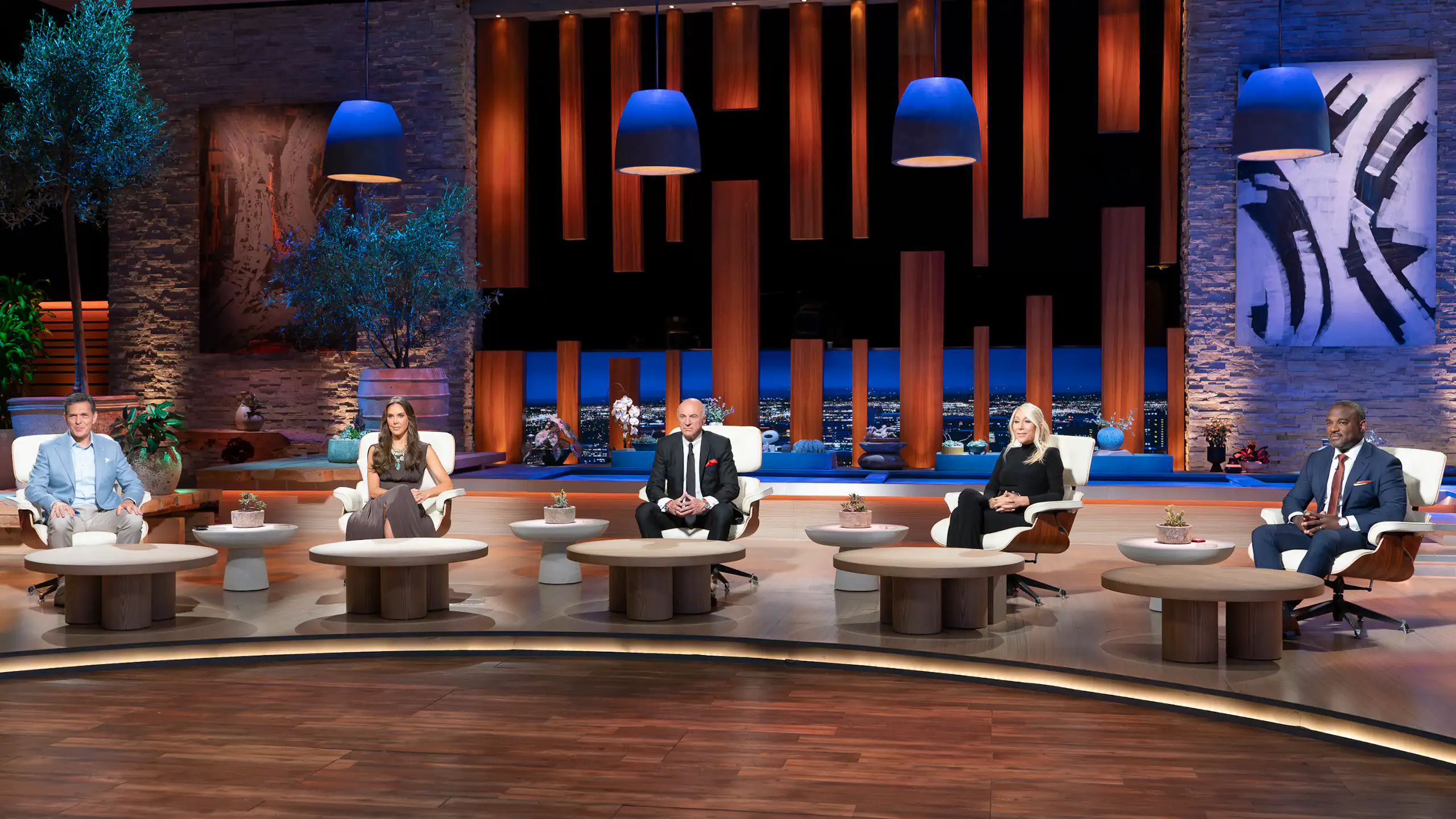 3 men and 2 women in formal clothes sit in chairs in front of tables on a big stage