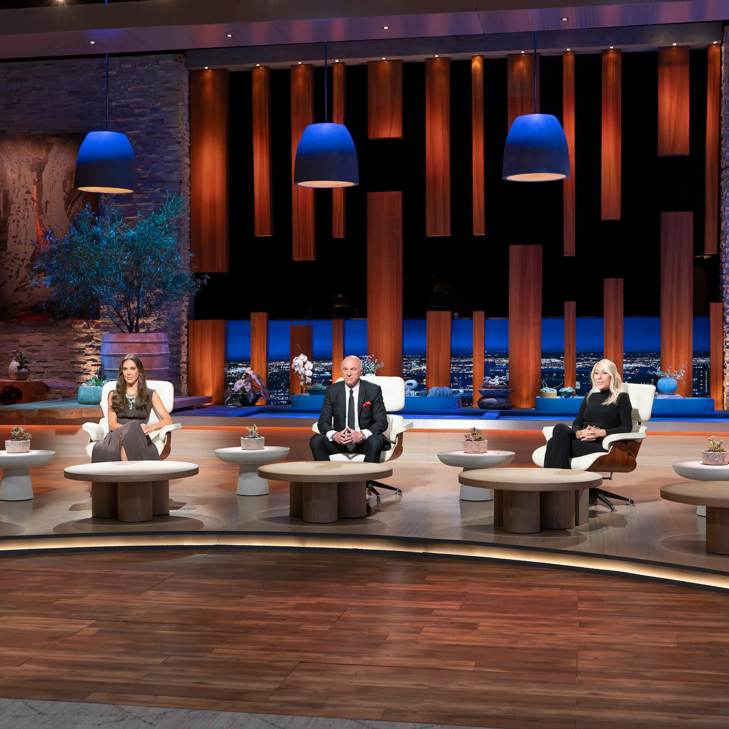 3 men and 2 women in formal clothes sit in chairs in front of tables on a big stage