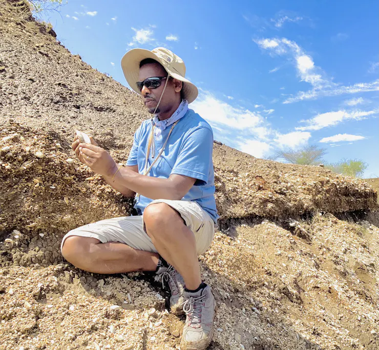 A scientist looks at a sample with layers of rock in the background.