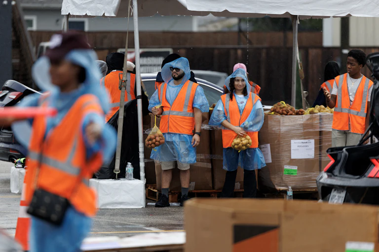 People in orange vests stand under tents with bags of produce.