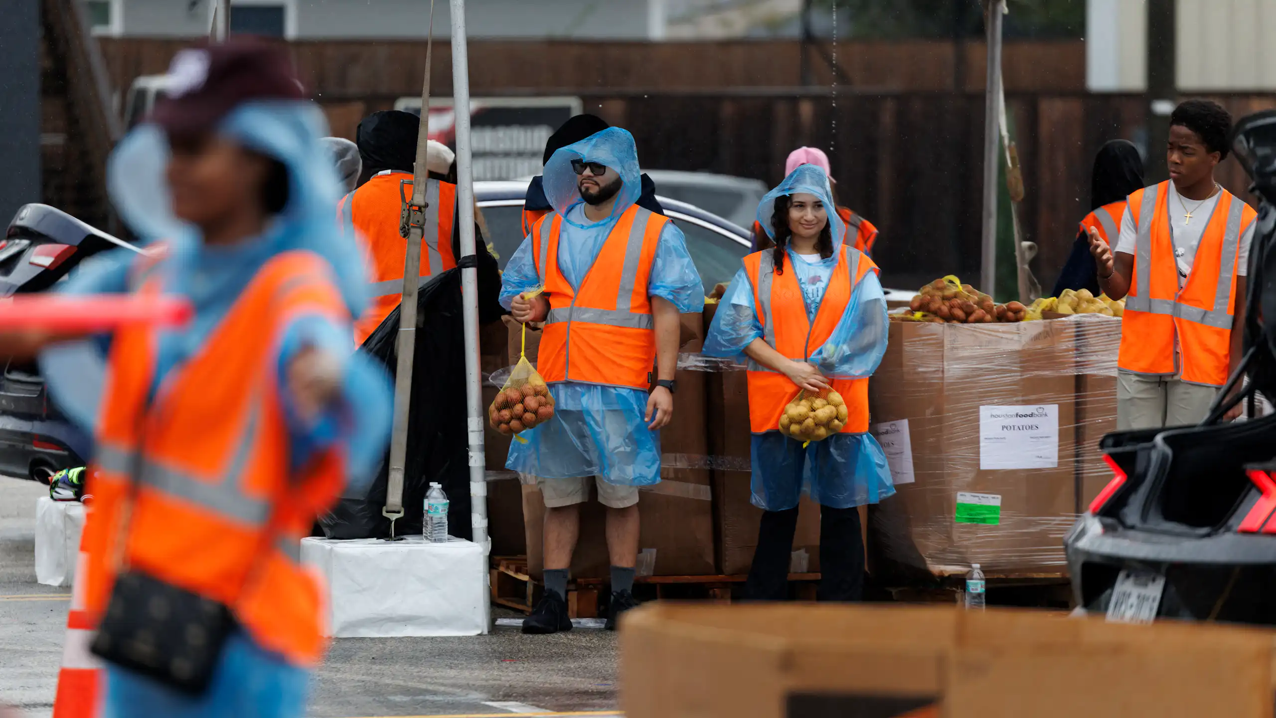 People in orange vests stand under tents with bags of produce.