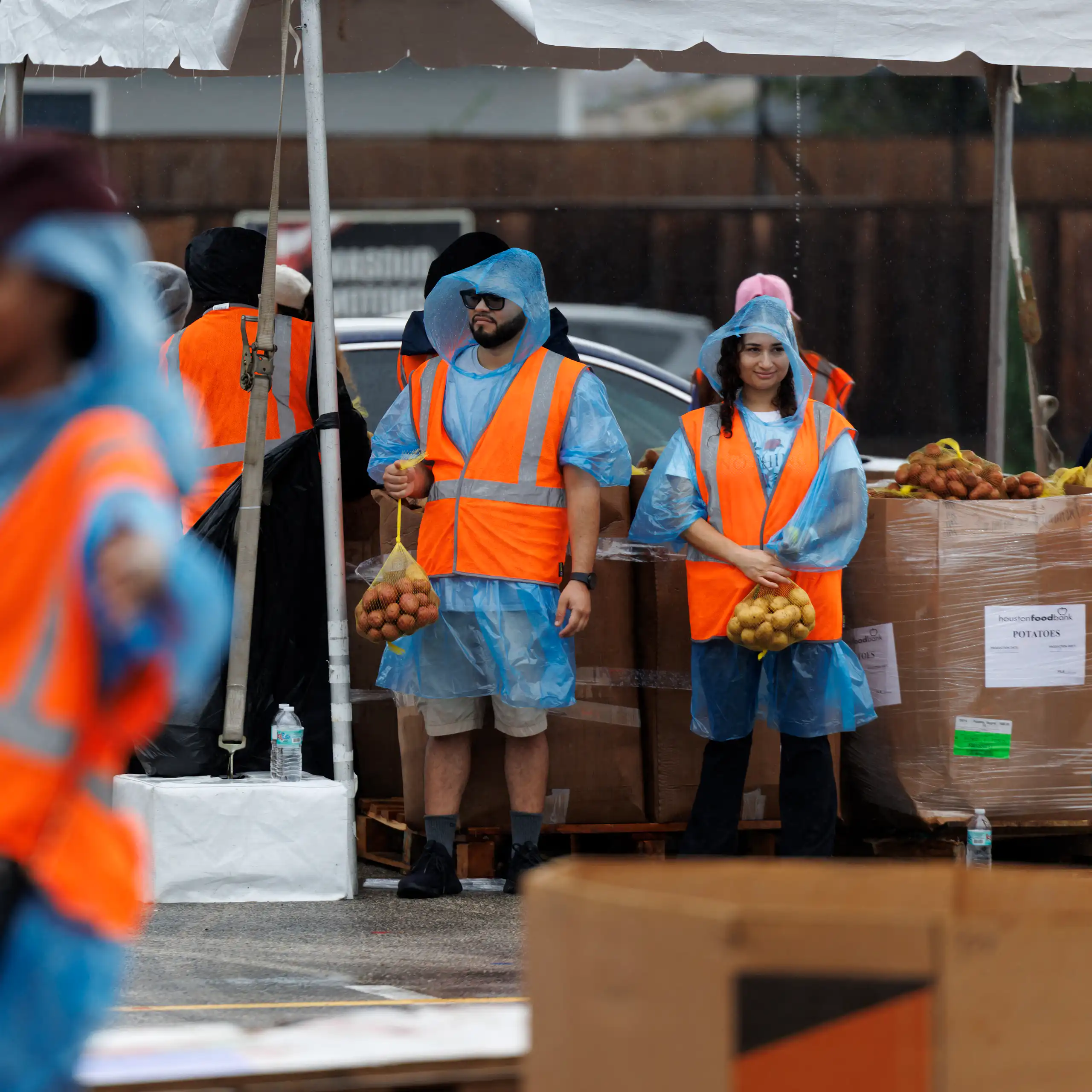 People in orange vests stand under tents with bags of produce.