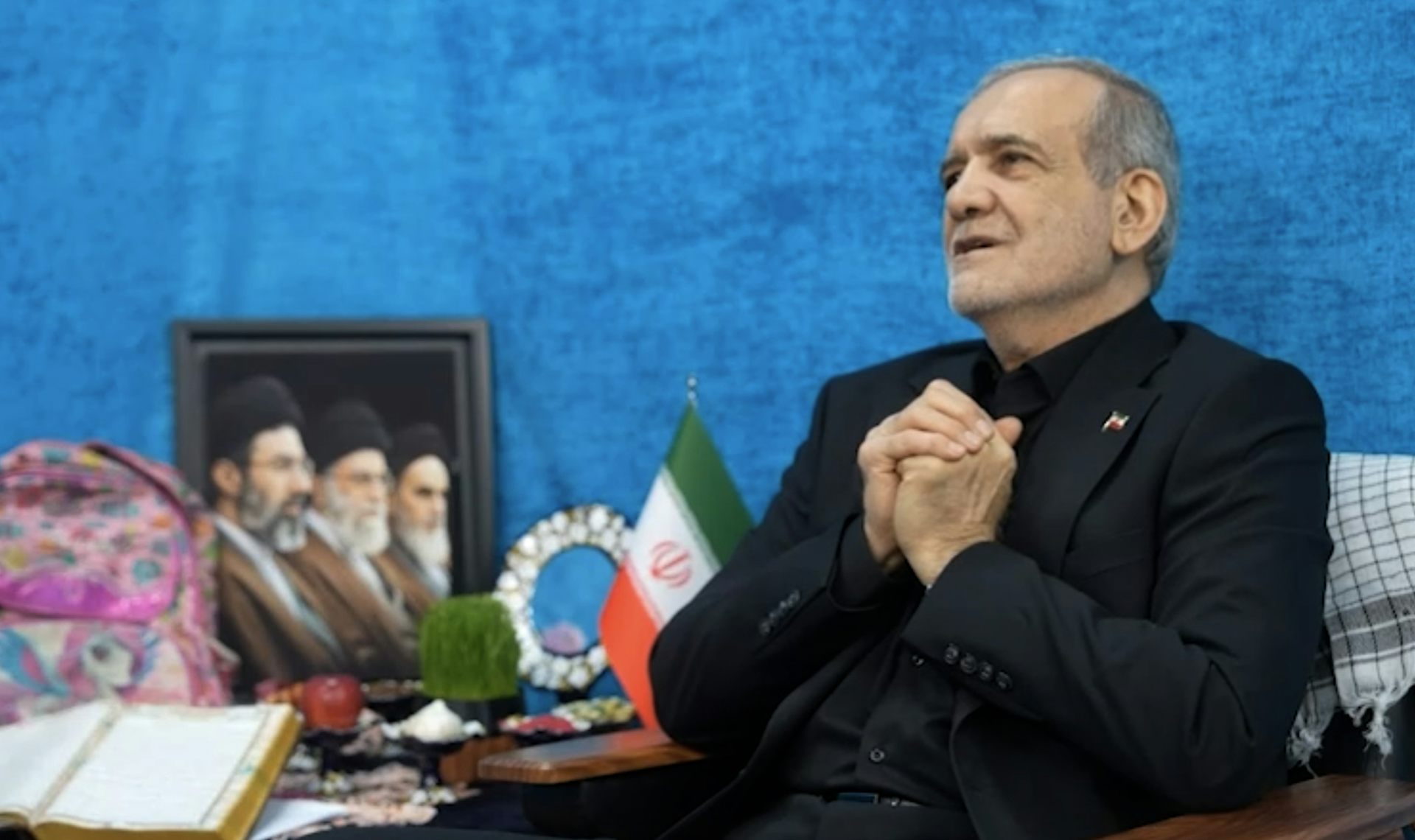 A man clasps his hands while sat in front of a photo of three men.