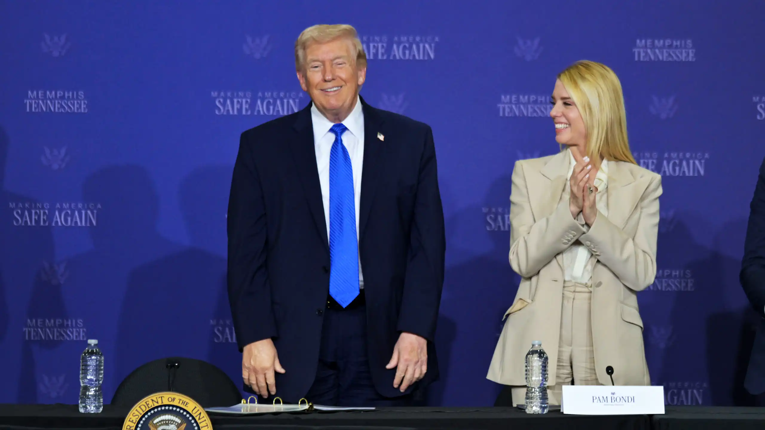 A man dressed in suit and tie stands on a stage next to a woman dressed in a beige suit.