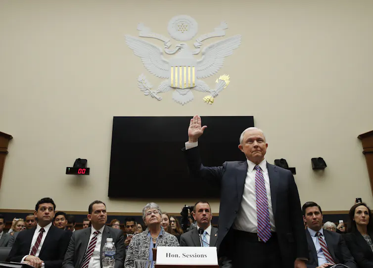 A man dressed in a suit and tie lifts his right hand in front of a panel of lawmakers.