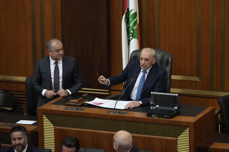 A men in a suit gestures from his speaker's chair.