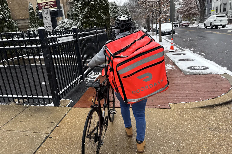 A DoorDash delivery person wears a large bag on their back as they walk with a bicycle on a snowy street.