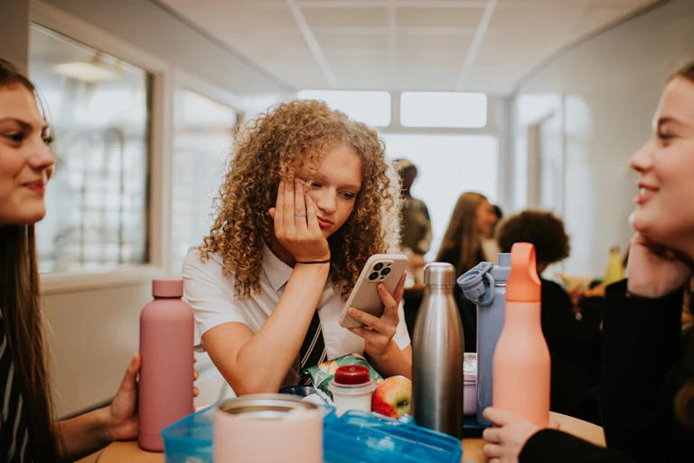 A teenage girl with holds a phone and looks at it, while two other teen girls sit around her at a table, with water bottles and food containers in front of them.
