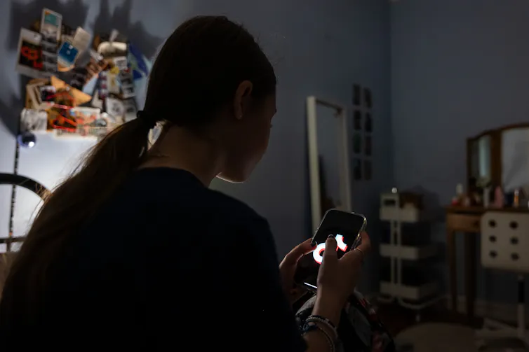 The outline of a teenage girl is seen in a dark room, as she looks at a phone in her hands.