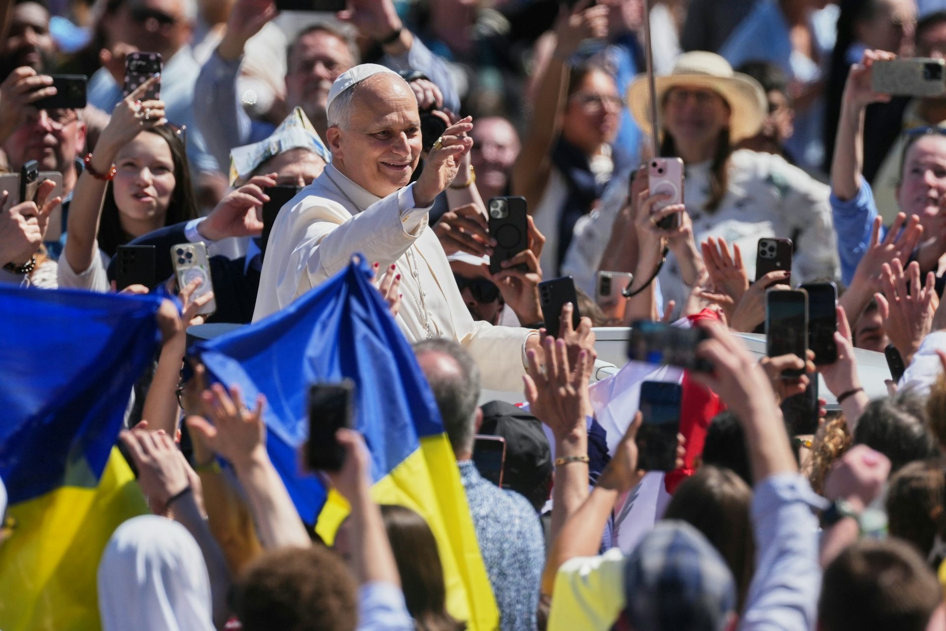 Un hombre con túnica blanca en medio de una multitud al aire libre sonríe en un día soleado. la gente ondea banderas ucranianas azules y amarillas.