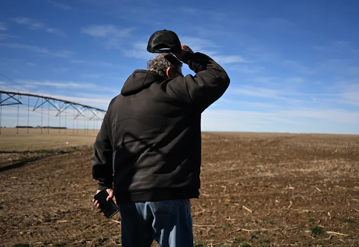 A man in a hoodie stands in a field, lifting his ballcap and scratching his head.
