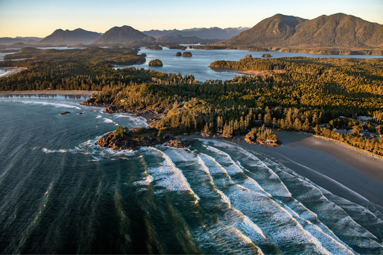 An aerial photo of a beach with trees and inlets and mountains in the background.
