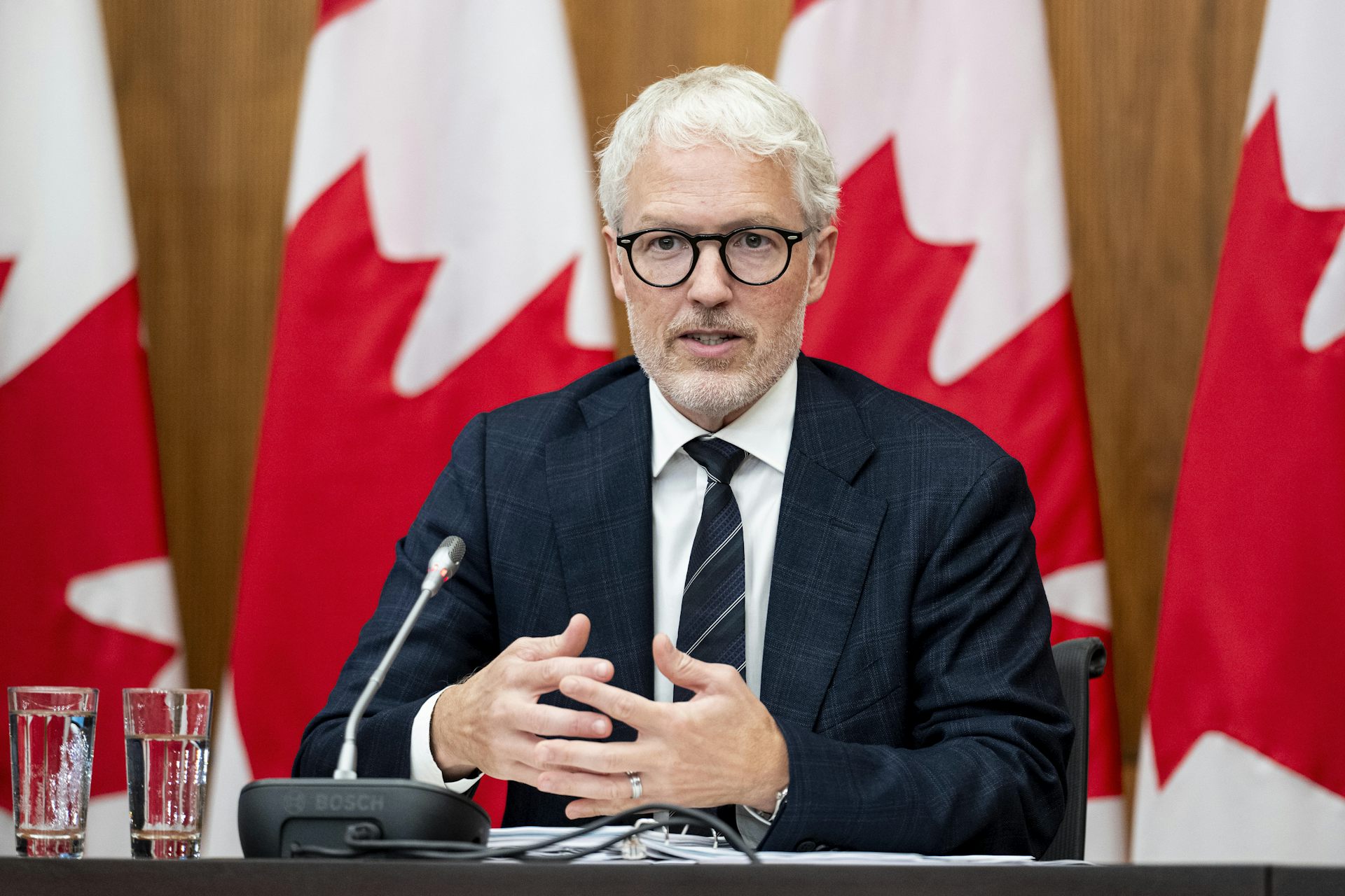 Un hombre blanco de mediana edad con pelo gris, barba y gafas habla sentado frente a una bandera canadiense