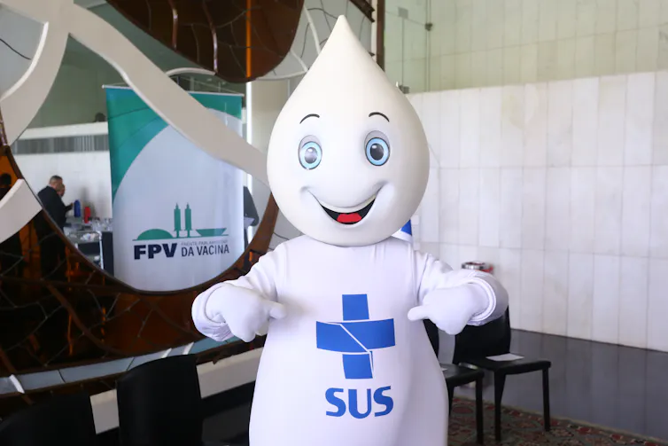 Uma gotinha de vacina branca com um rosto sorridente e o logotipo do sistema público de saúde do Brasil na barriga.