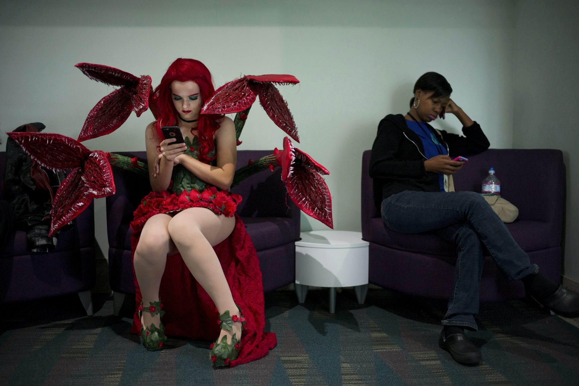 Una persona vestida de rojo y verde con tentáculos parecidos a plantas, tacones altos y lápiz labial, se sienta en una silla y mira el teléfono.