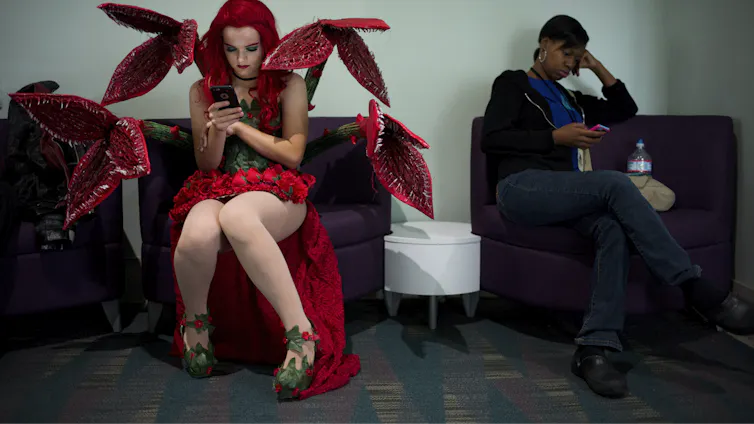 A person in a red and green costume with plant-like tentacles, wearing high heels and lipstick, sits in a chair looking at a phone.