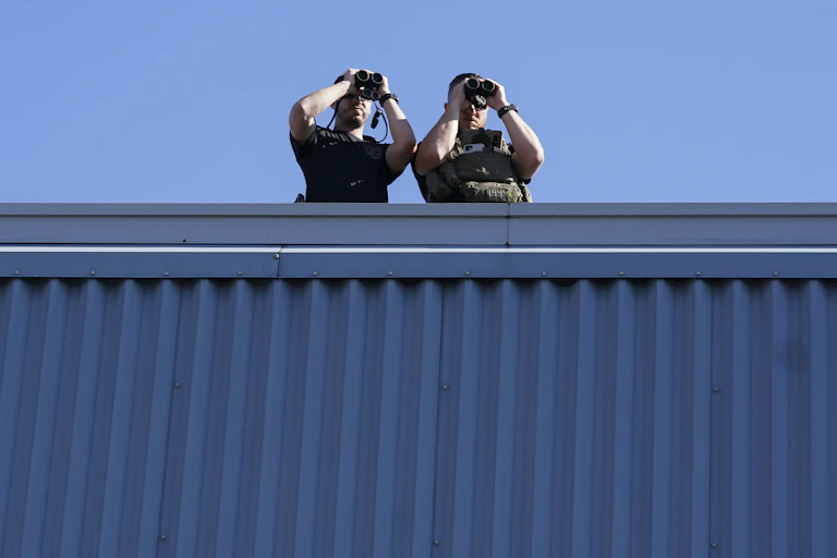 Two armed police officers stand on a roof, both looking through binoculars.