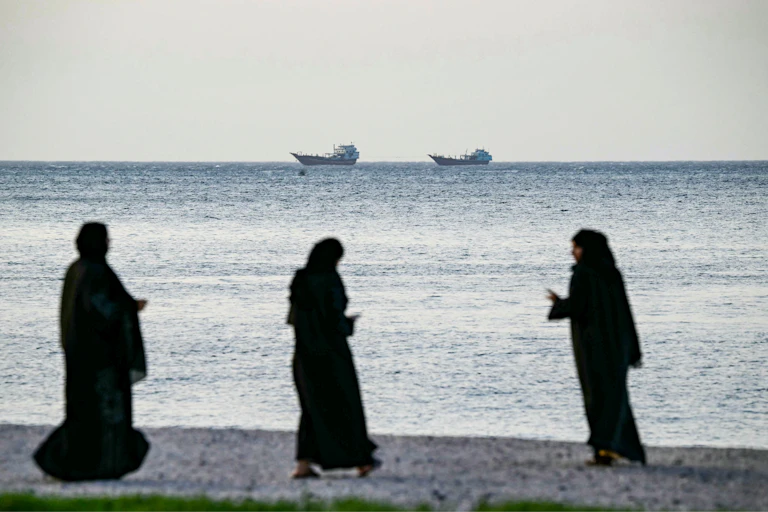Women dressed in black burqas walk on the beach in front of the ocean with two large ships sailing on the horizon