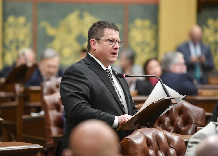 A middle-aged man with glasses speaks at a lectern.