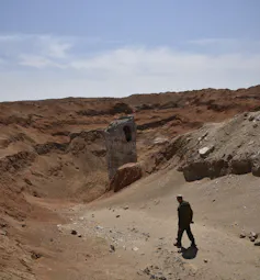 La silueta de un hombre camina en primer plano de un paisaje polvoriento con un gran objeto metálico que sobresale de un cráter.