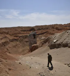 silhouetted man walks in foreground of dusty landscape with a large metallic object poking out of crater