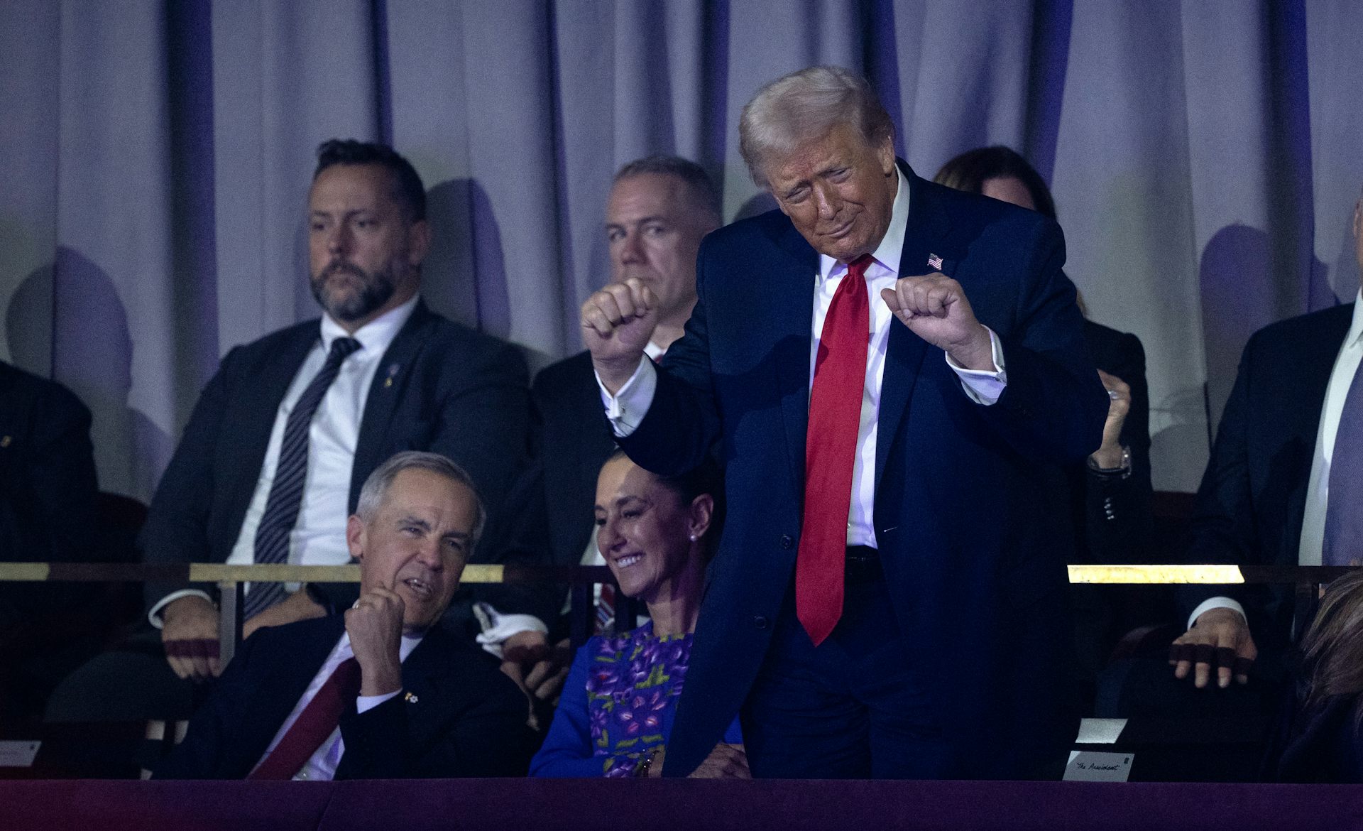 Un hombre anciano y corpulento con el pelo esponjoso de color rubio canoso baila torpemente mientras un hombre y una mujer se sientan a su lado y conversan.