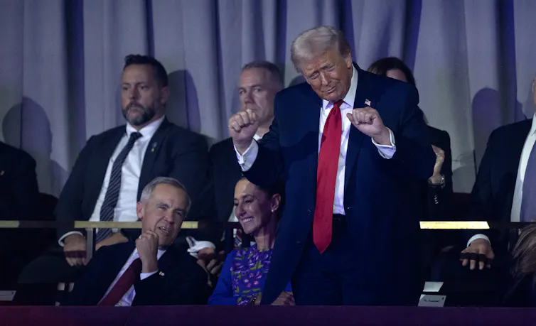 Un hombre anciano y corpulento con el pelo esponjoso de color rubio canoso baila torpemente mientras un hombre y una mujer se sientan a su lado y conversan.