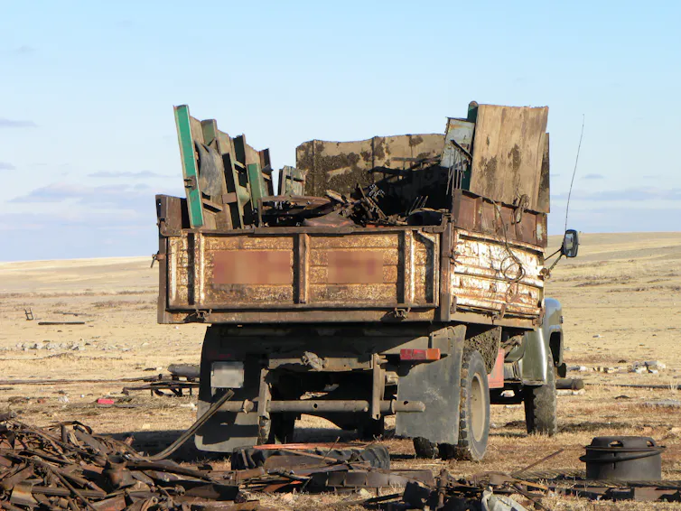 Un camión con metal amontonado en la parte trasera, estacionado en un paisaje abierto, metal recuperado en el suelo a su alrededor.