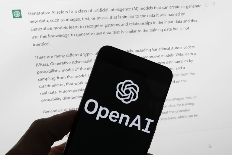 The OpenAI logo is displayed on a mobile phone in front of a computer screen showing text output from ChatGPT.