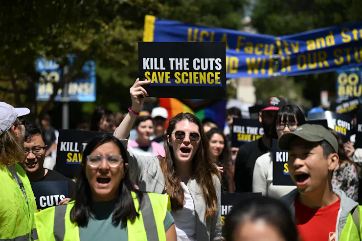 A group of young people stand together and hold signs outside. Some of the people wear neon yellow vests. One of the signs says Kill the cuts save science!