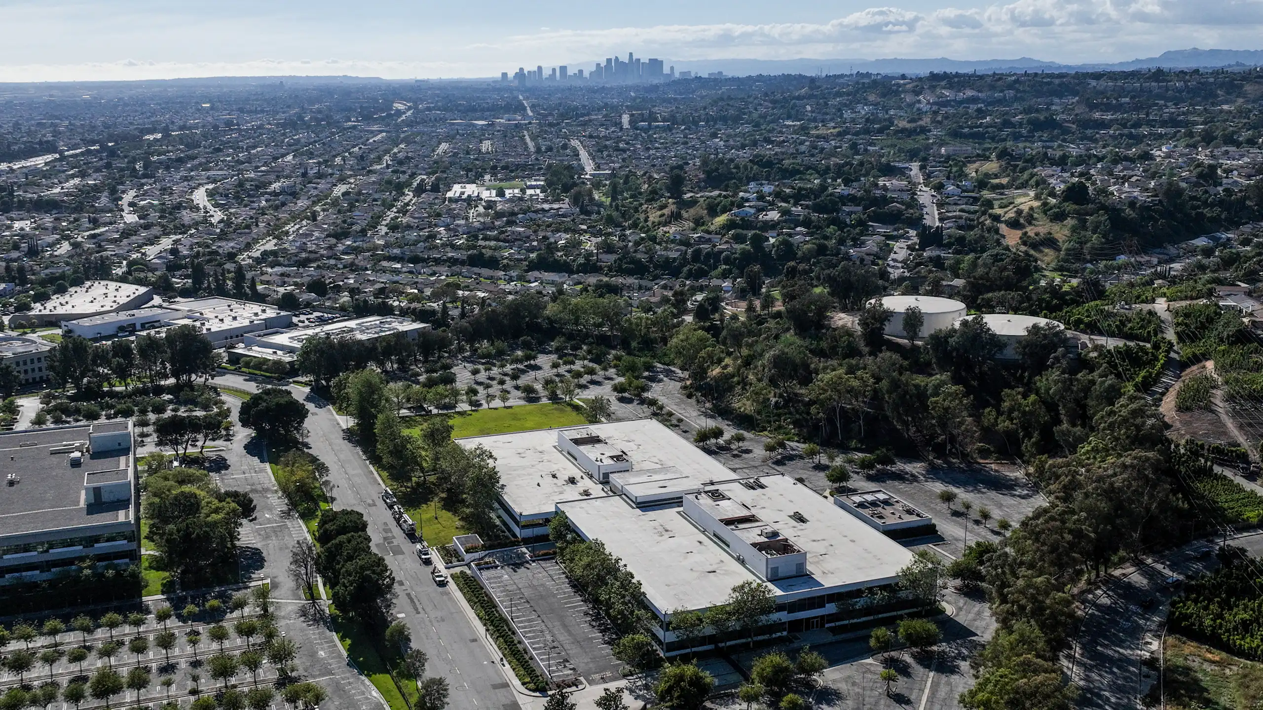 An aerial view shows a large building in the foreground with homes and businesses nearby, and skyscrapers in the distance.