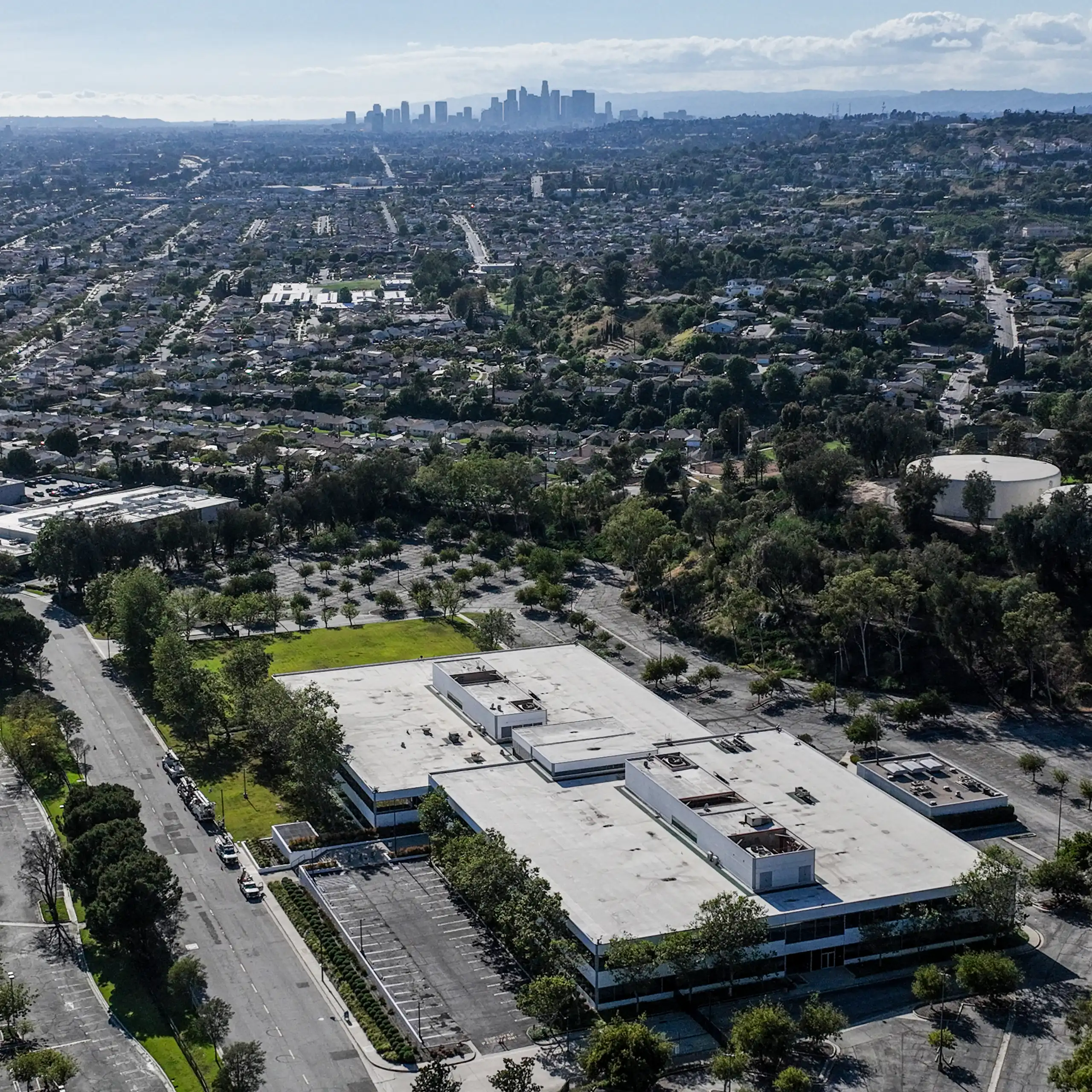 An aerial view shows a large building in the foreground with homes and businesses nearby, and skyscrapers in the distance.