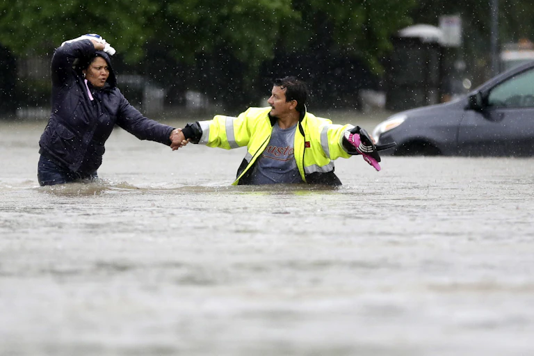 A man in a reflective jacket reaches for a woman's hand as they try to walk through waist-deep water. A car behind them is in water up to its windows.