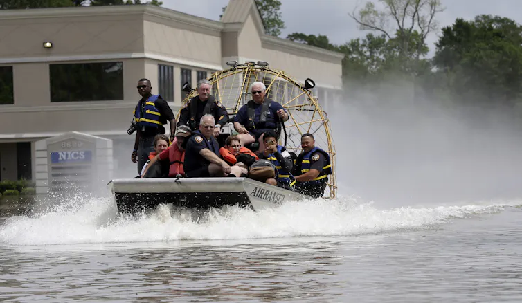 People in an airboat going past buildings surrounded by water.