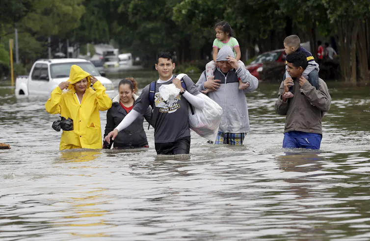 ¿Qué pasaría si la destructiva inundación del Día de Impuestos de Texas se centrara en el interior de Houston? Por eso las ciudades necesitan planificar para lo increíble 1 La gente carga sus pertenencias en bolsas de basura y los adultos llevan a niños pequeños sobre sus hombros mientras caminan por el agua que les llega hasta la cintura.