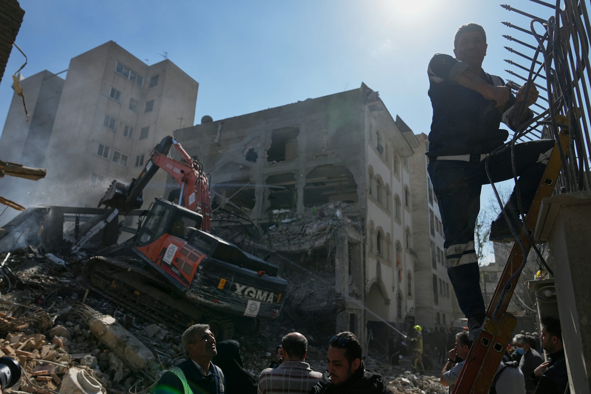 A fireman on a ladder in the foreground with destroyed buildings and a bulldozer behind him.