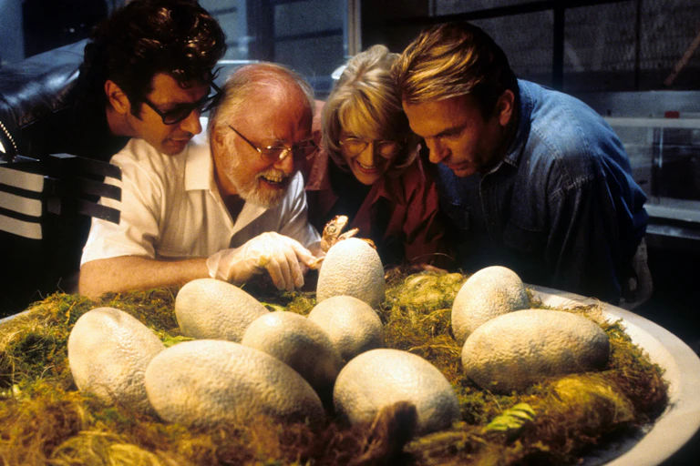 Imagem do filme “Jurassic Park” mostrando quatro pessoas examinando um ninho de ovos grandes e de aparência coriácea.