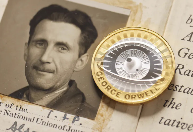 A gold-colored coin with an engraving reading 'Big Brother is Watching You' lies next to a sepia-toned portrait of a man with a thin mustache.
