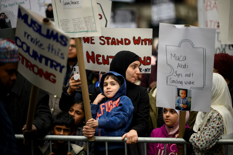 A woman wearing a headscarf stands with a child at a protest, holding a sign that reads, 'Muslims make this country strong.'