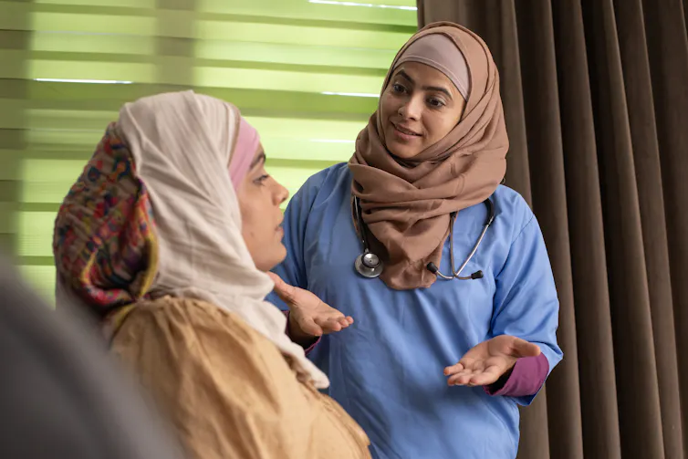 A woman wearing a headscarf speaks with another woman reclining on a bed, who is also wearing a headscarf.
