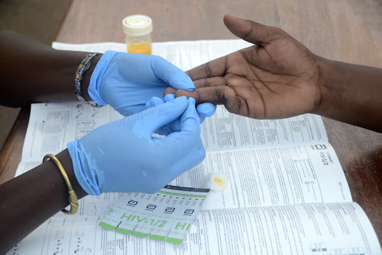 Hands in blue gloves hold another person's hand, pricking a finger