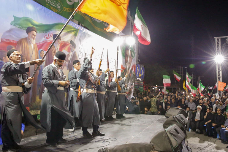Iranian pro-governmentrally with traditionally dressed supporters holding rifles on a stage backed by patriotic images.
