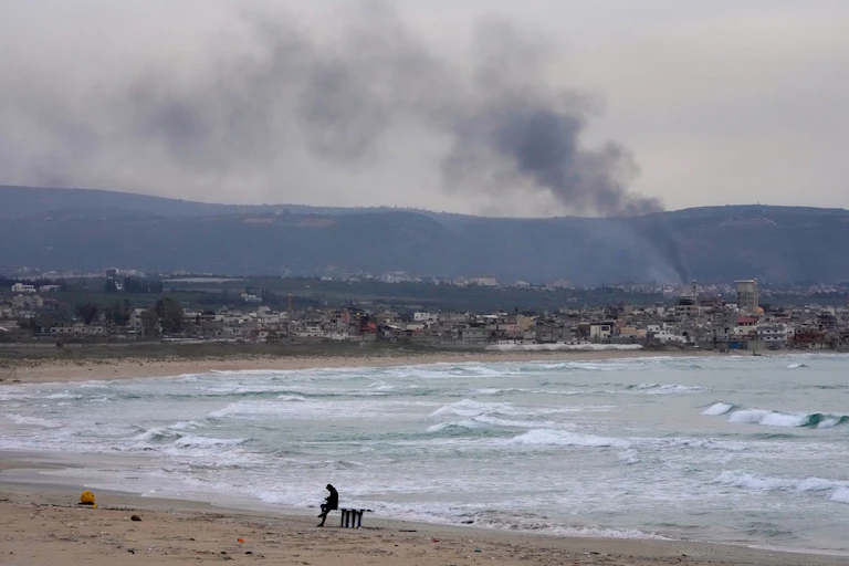 A man sits next to the ocean as smoke billows in the background.