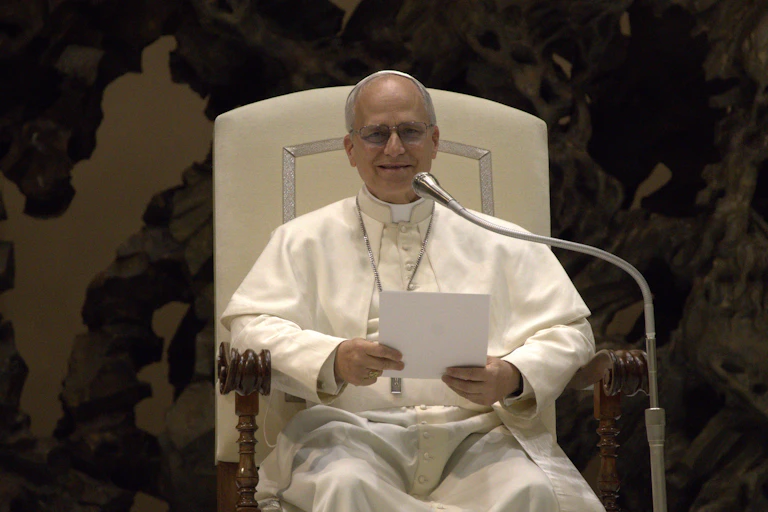 A seated man dressed in white robes holding a piece of white paper and speaking into a microphone